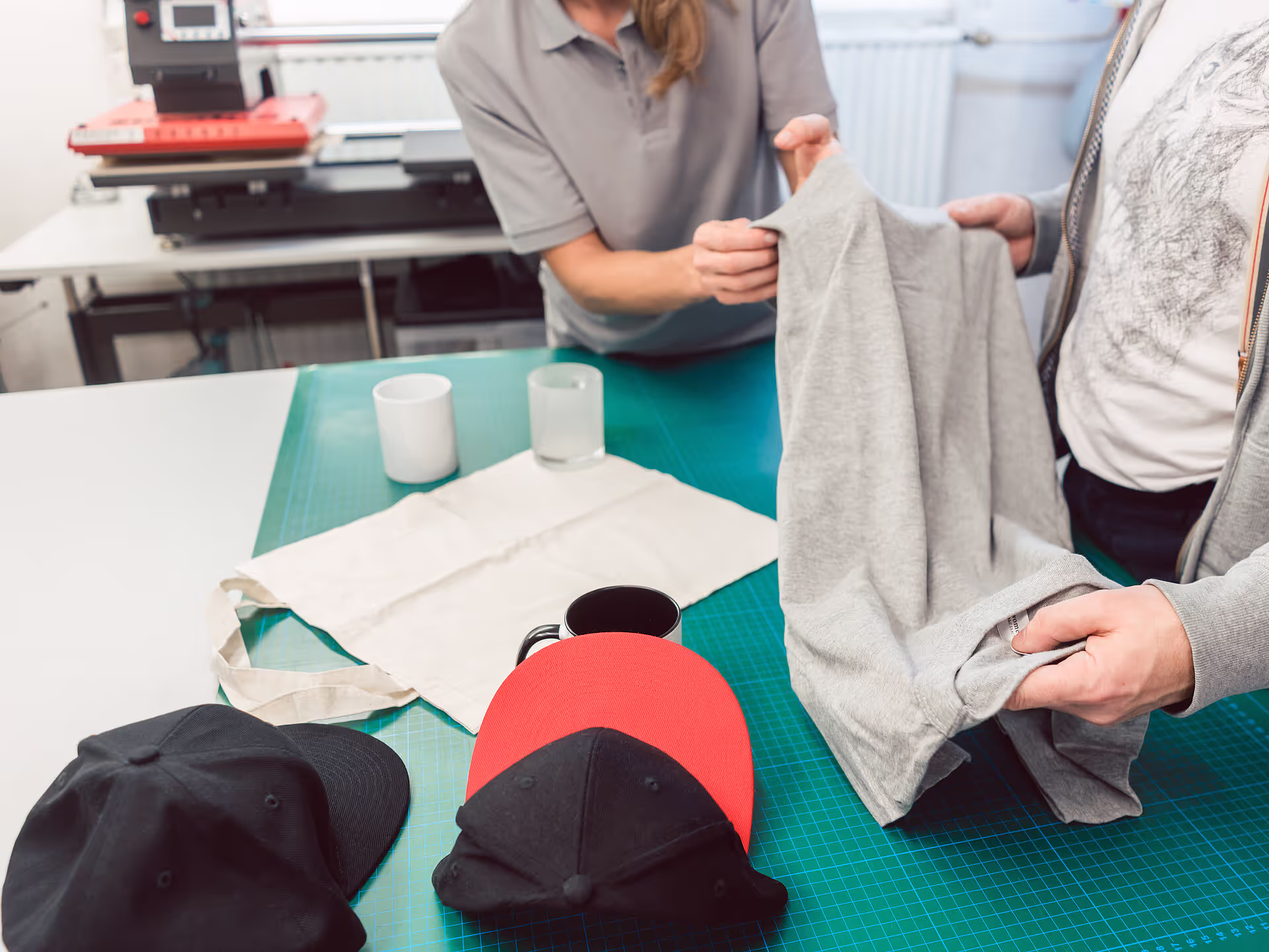Zwei Personen begutachten ein graues T-Shirt auf einem Tisch mit leeren Tassen, Tragetaschen und Baseballkappen, was auf einen Arbeitsbereich für den individuellen Druck oder die Herstellung von Waren hindeutet. Im Hintergrund ist eine Druckmaschine zu sehen.