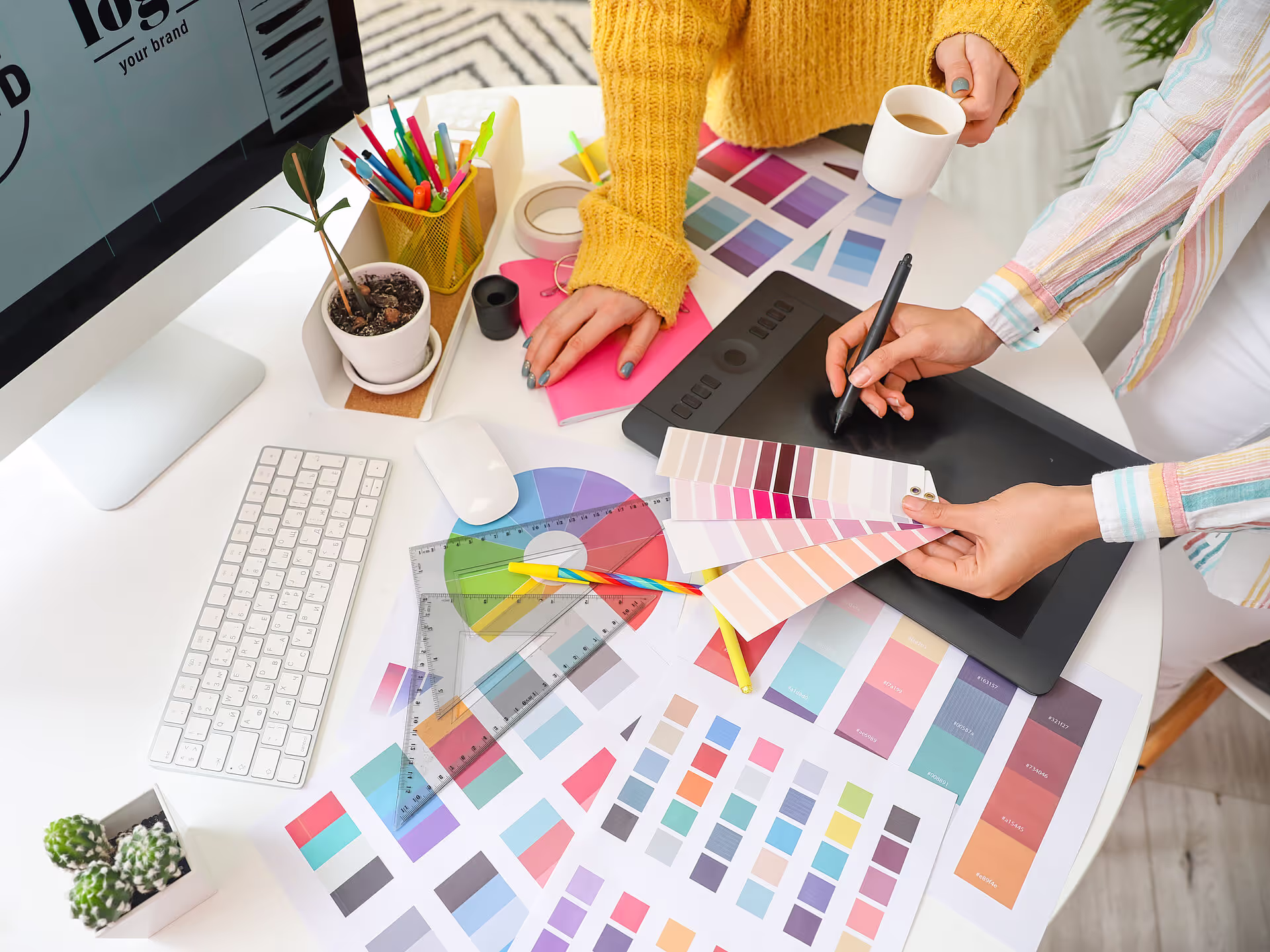 Zwei Personen arbeiten an einem Schreibtisch, der mit Farbmustern, Diagrammen, einer Tastatur und Designwerkzeugen bedeckt ist; eine verwendet ein Stifttablett, während die andere Farbmuster hält. Ein heller, kreativer Arbeitsbereich mit sichtbaren Pflanzen und Materialien.