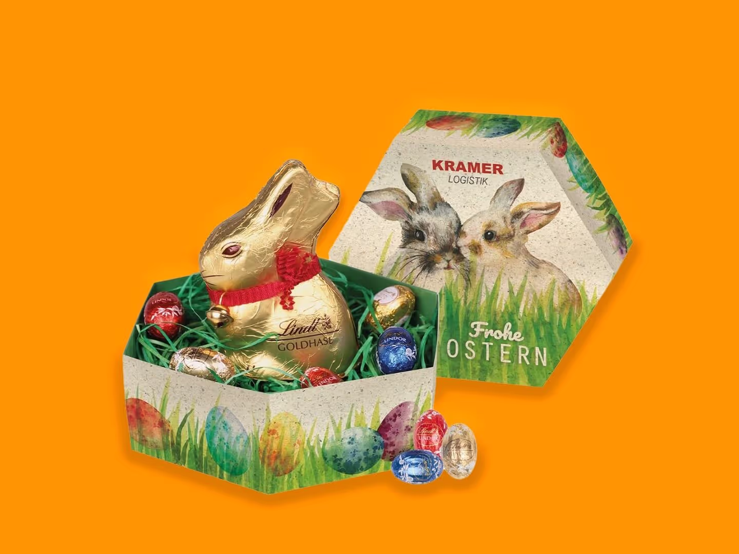 Eine sechseckige Ostergeschenkbox mit bemalten Eiern und Hasen enthält einen in Gold verpackten Schokoladenhasen und verschiedene bunte Schokoladeneier. Der Deckel, auf dem zwei Hasen abgebildet sind, ruht neben der Schachtel auf einem orangefarbenen Hintergrund.